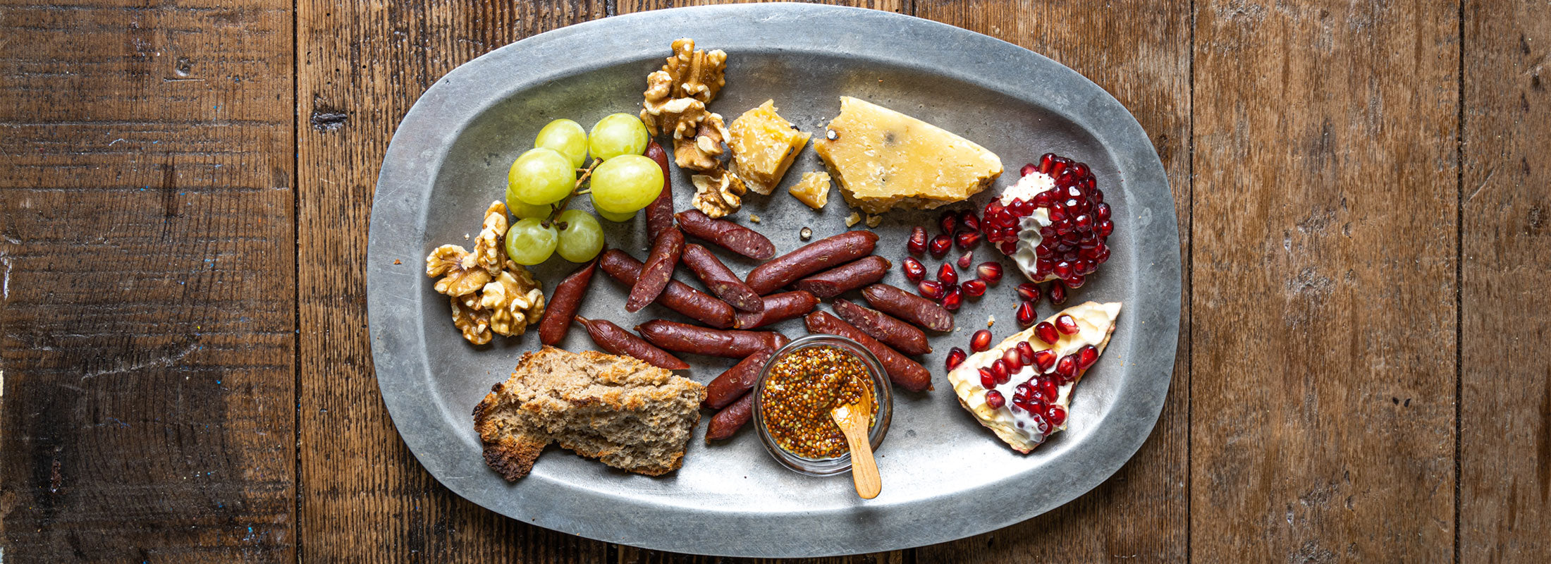 A metal tray on a wooden table serves Patagonia Provisions Buffalo Links, cheeses, fruits, and bread